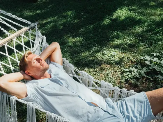 Mann i lyseblå skjorte og shorts slapper av og nyter solen i en hengekøye