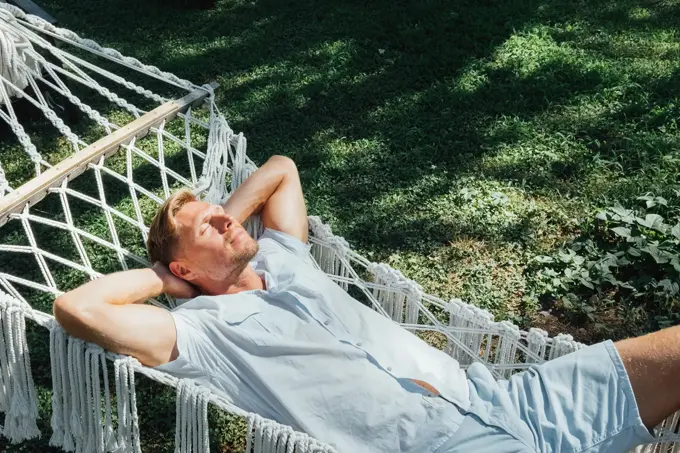 Mann i lyseblå skjorte og shorts slapper av og nyter solen i en hengekøye