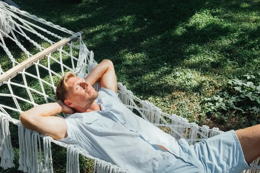 Mann i lyseblå skjorte og shorts slapper av og nyter solen i en hengekøye