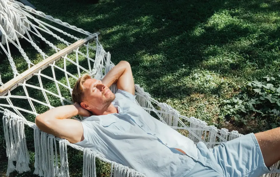 Mann i lyseblå skjorte og shorts slapper av og nyter solen i en hengekøye