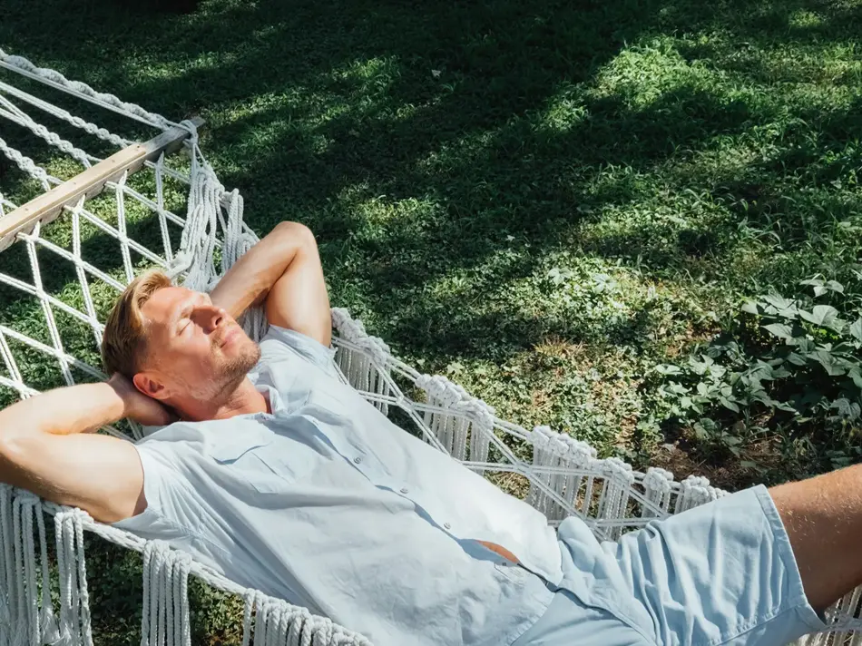 Mann i lyseblå skjorte og shorts slapper av og nyter solen i en hengekøye