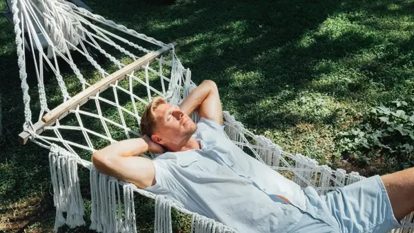 Mann i lyseblå skjorte og shorts slapper av og nyter solen i en hengekøye