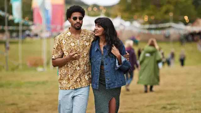 Et ungt par på festival. Mannen er iført mønstrete skjorte og solbriller. Kvinnen har denimjakke. Mannen holder armen rundt kvinnen. 