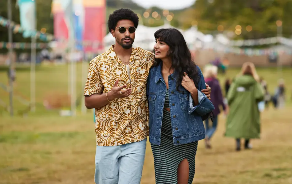 Et ungt par på festival. Mannen er iført mønstrete skjorte og solbriller. Kvinnen har denimjakke. Mannen holder armen rundt kvinnen. 