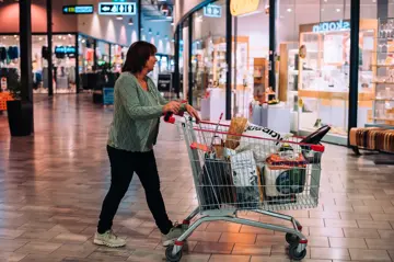 Bildet er fra kjøpesenteret Charlottenbergs Shoppingcenter og viser en dame i grønn strikkejakke med en handlekurv full av ulike bæreposer.