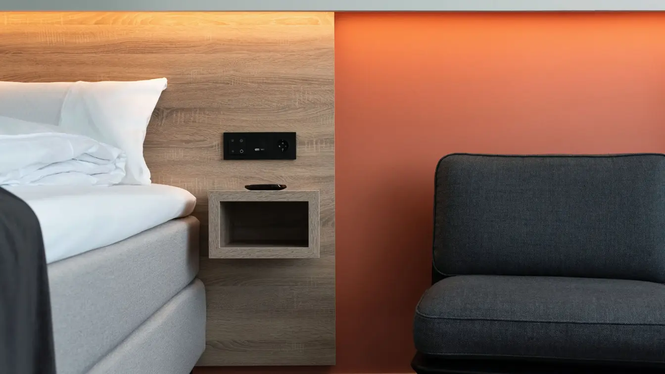Detaljbilde av sengegavl i træ, lite vegghent nattbord, oransje vegg og grå stol.  I business rom på Thon Hotel Norge i Kristiansand