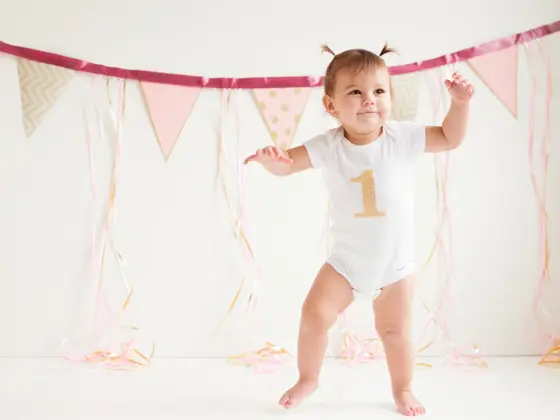 A baby girl is walking her first steps. One foot is on the floor and the other one is moving. She has a beautiful smile on her face. She has two ponytails on her head. She is wearing a onsie with the number one printed on it. It’s her birthday. There’s three balloons beside her side and decoration in the background.