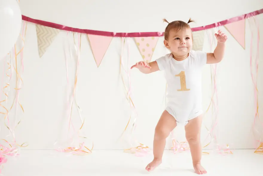 A baby girl is walking her first steps. One foot is on the floor and the other one is moving. She has a beautiful smile on her face. She has two ponytails on her head. She is wearing a onsie with the number one printed on it. It’s her birthday. There’s three balloons beside her side and decoration in the background.