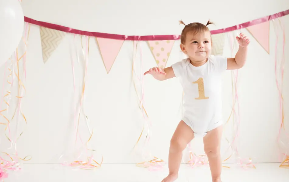 A baby girl is walking her first steps. One foot is on the floor and the other one is moving. She has a beautiful smile on her face. She has two ponytails on her head. She is wearing a onsie with the number one printed on it. It’s her birthday. There’s three balloons beside her side and decoration in the background.