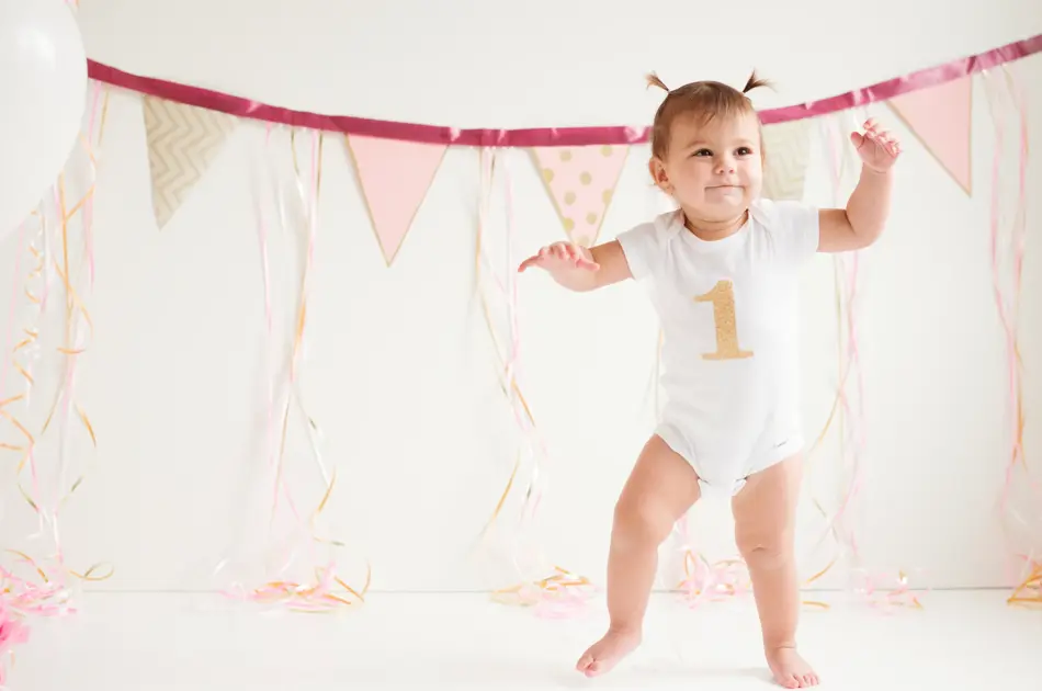 A baby girl is walking her first steps. One foot is on the floor and the other one is moving. She has a beautiful smile on her face. She has two ponytails on her head. She is wearing a onsie with the number one printed on it. It’s her birthday. There’s three balloons beside her side and decoration in the background.