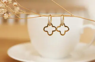 Et par øreringer med kløverdesign henger på en hvit blomst foran en kaffekopp.