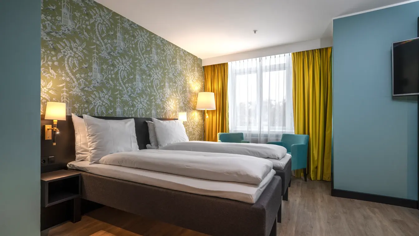 Dobbeltseng med hvitt sengetøy, gull gardiner i bakgrunnen med turkise stoler plassert foran. Mønstret tapet og parkett gulv.