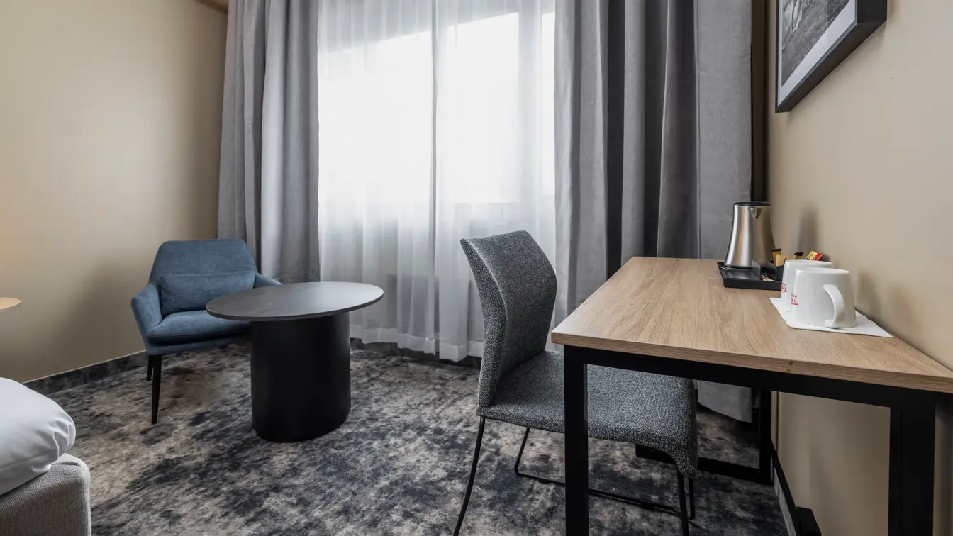 Dobbeltrom med lenestol og tilhørende bord, skrivebord med stol, lange gardiner og stort vindu.