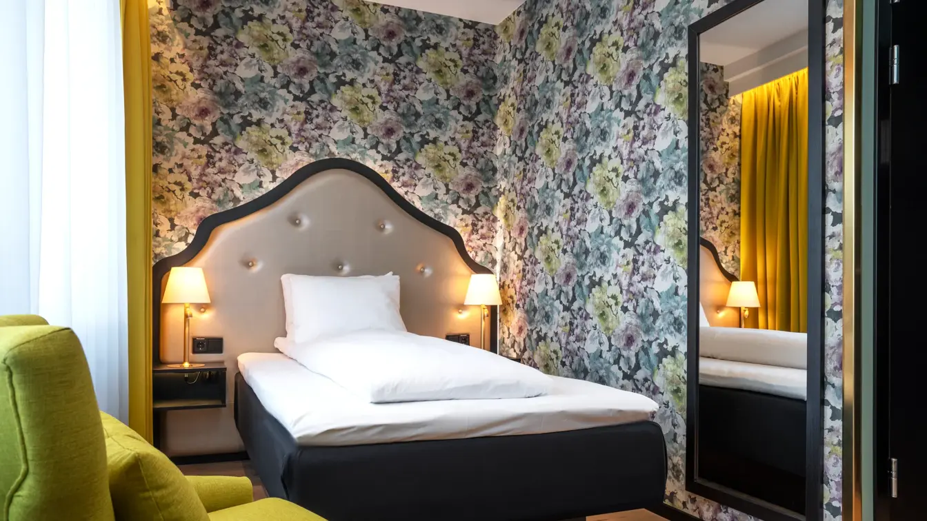 Gul stol, queen seng med sengegavl, blomstrete tapet og stort vegghengt speil i standard rom queen på Thon Hotel Cecil.