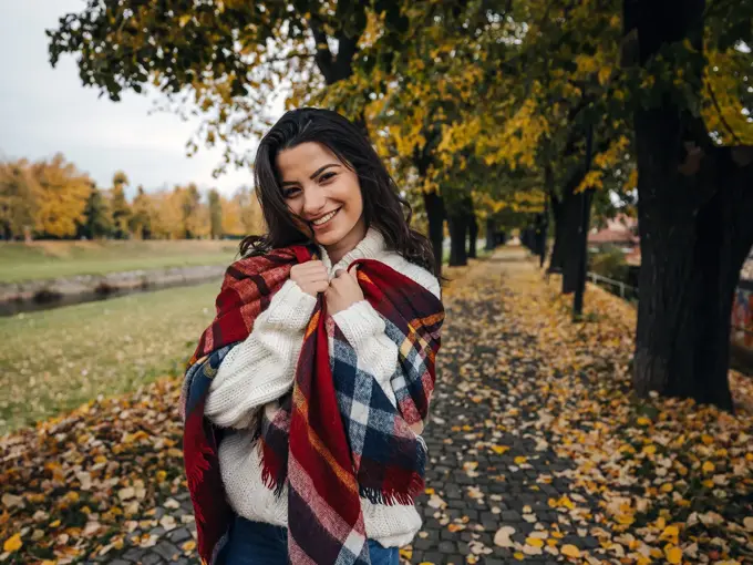 Dame kledd i hvit ullgenser og stort ullsjal med blåde og røde ruter står i en park og smiler mot kamera.