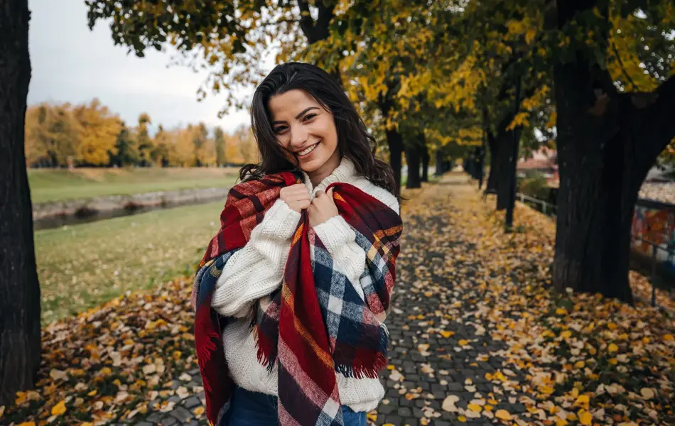 Dame kledd i hvit ullgenser og stort ullsjal med blåde og røde ruter står i en park og smiler mot kamera.
