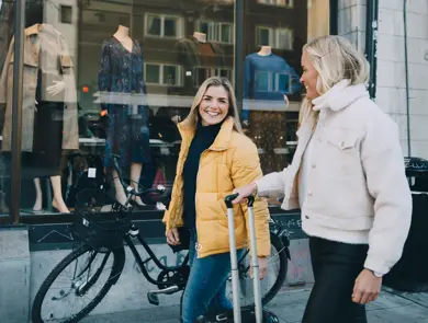 En dame med sykkel og en gul boblejakke og en dame med en fluffy jakke i lyse farger og trillekoffert går gjennom et bymiljø