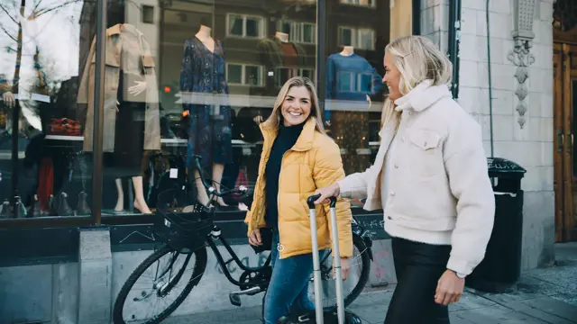 En dame med sykkel og en gul boblejakke og en dame med en fluffy jakke i lyse farger og trillekoffert går gjennom et bymiljø
