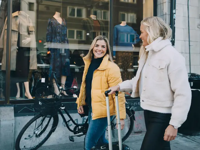 En dame med sykkel og en gul boblejakke og en dame med en fluffy jakke i lyse farger og trillekoffert går gjennom et bymiljø