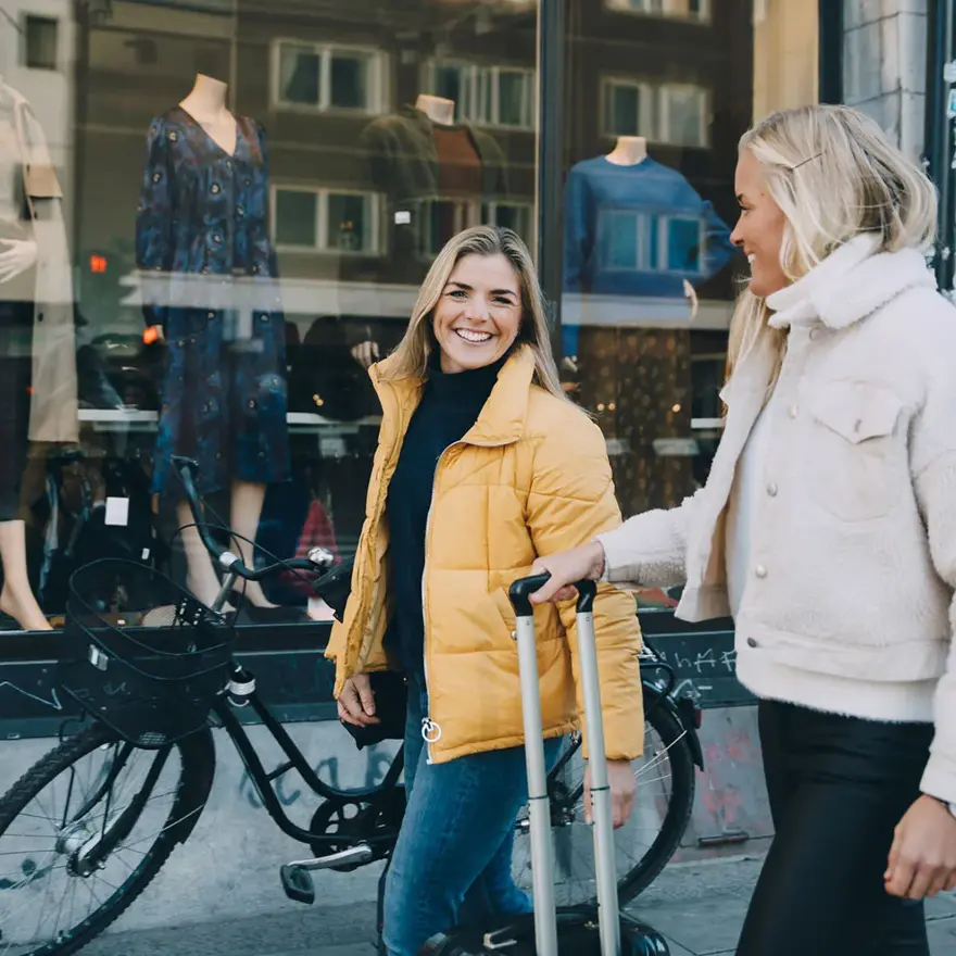 En dame med sykkel og en gul boblejakke og en dame med en fluffy jakke i lyse farger og trillekoffert går gjennom et bymiljø
