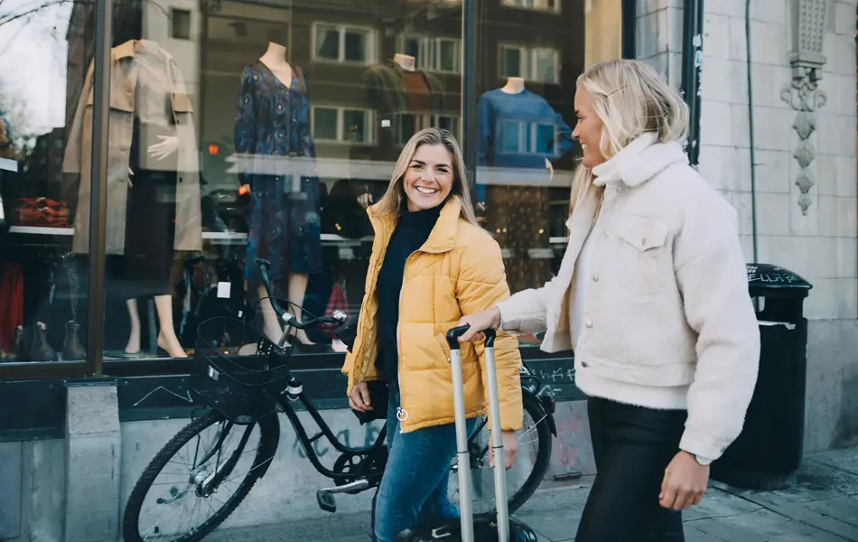En dame med sykkel og en gul boblejakke og en dame med en fluffy jakke i lyse farger og trillekoffert går gjennom et bymiljø