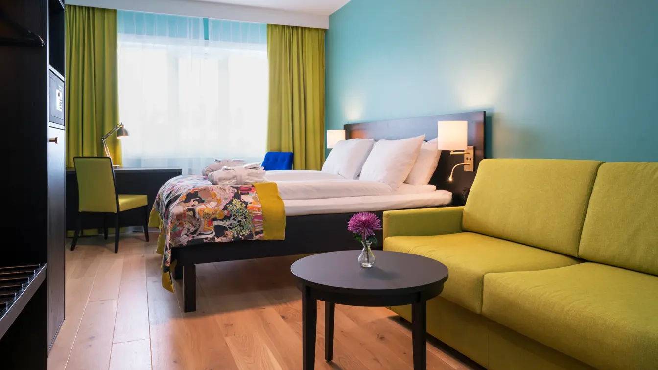 Gul sofa, bord, garderobeskap, dobbeltseng, skrivebord og lenestol på Buisness room på Thon Hotel Europa i Oslo sentrum rett ved Slottsparken