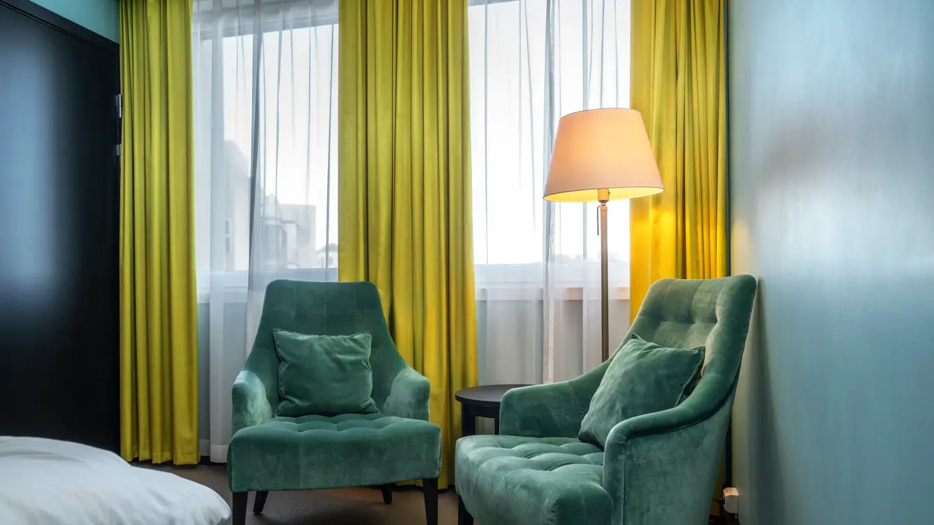 Dobbeltrom med to grønne velour lenestoler, stålampe og gule gardiner.
