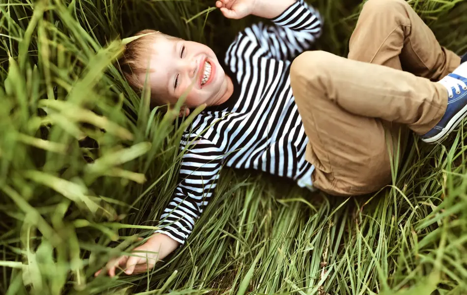 Gutt ler og ligger i gresset iført stripete genser og beige bukse