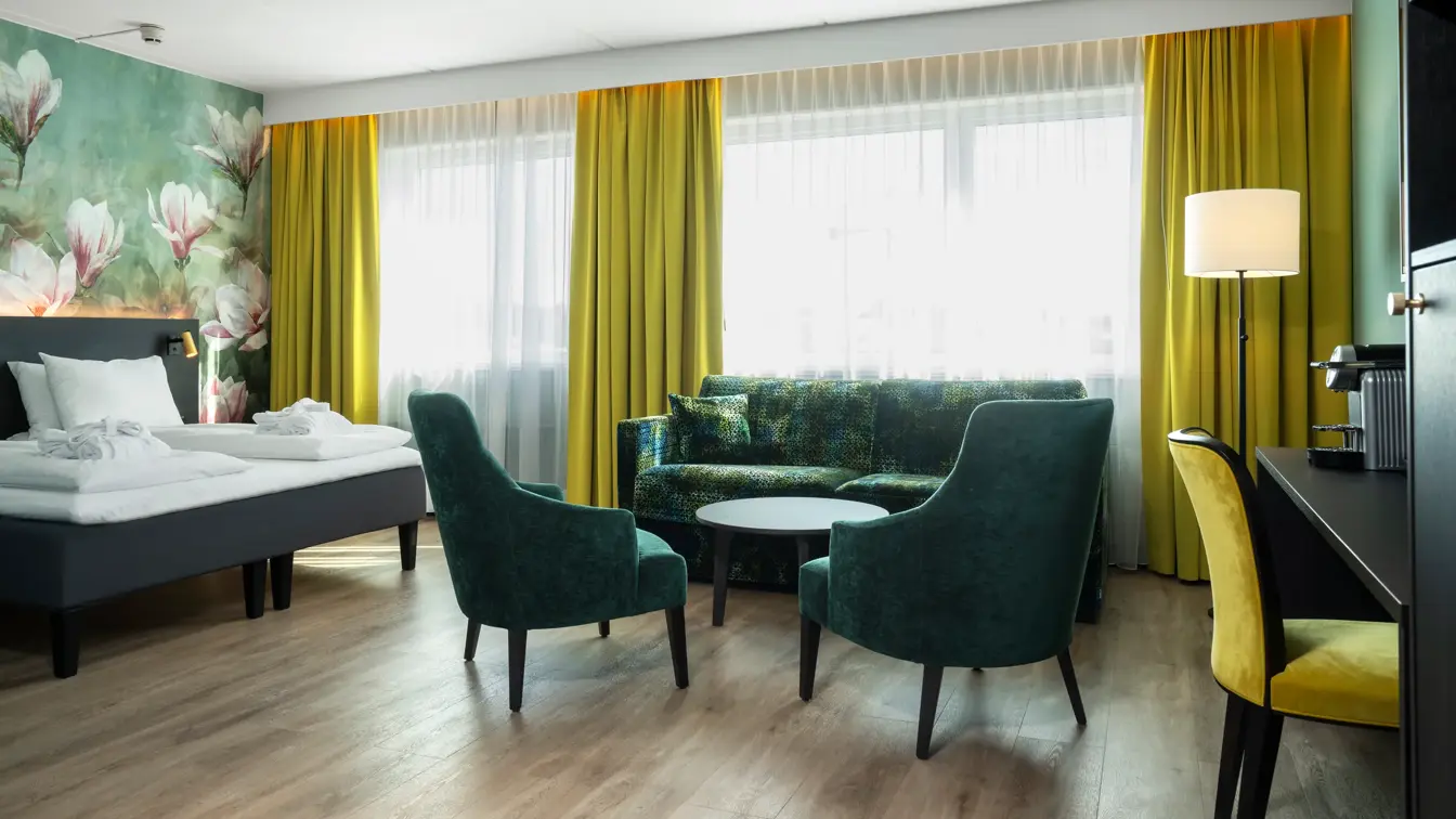 Dobbeltseng, blomstertapetvegg, vindu, gule gardiner, sofa, to grønne lenestoler, skrivebord, gul lenestol, kaffemaskin