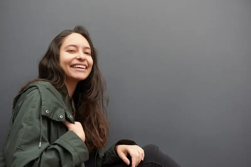 Dame med olivengrønn jakke poserer smilende sittende foran grå vegg 