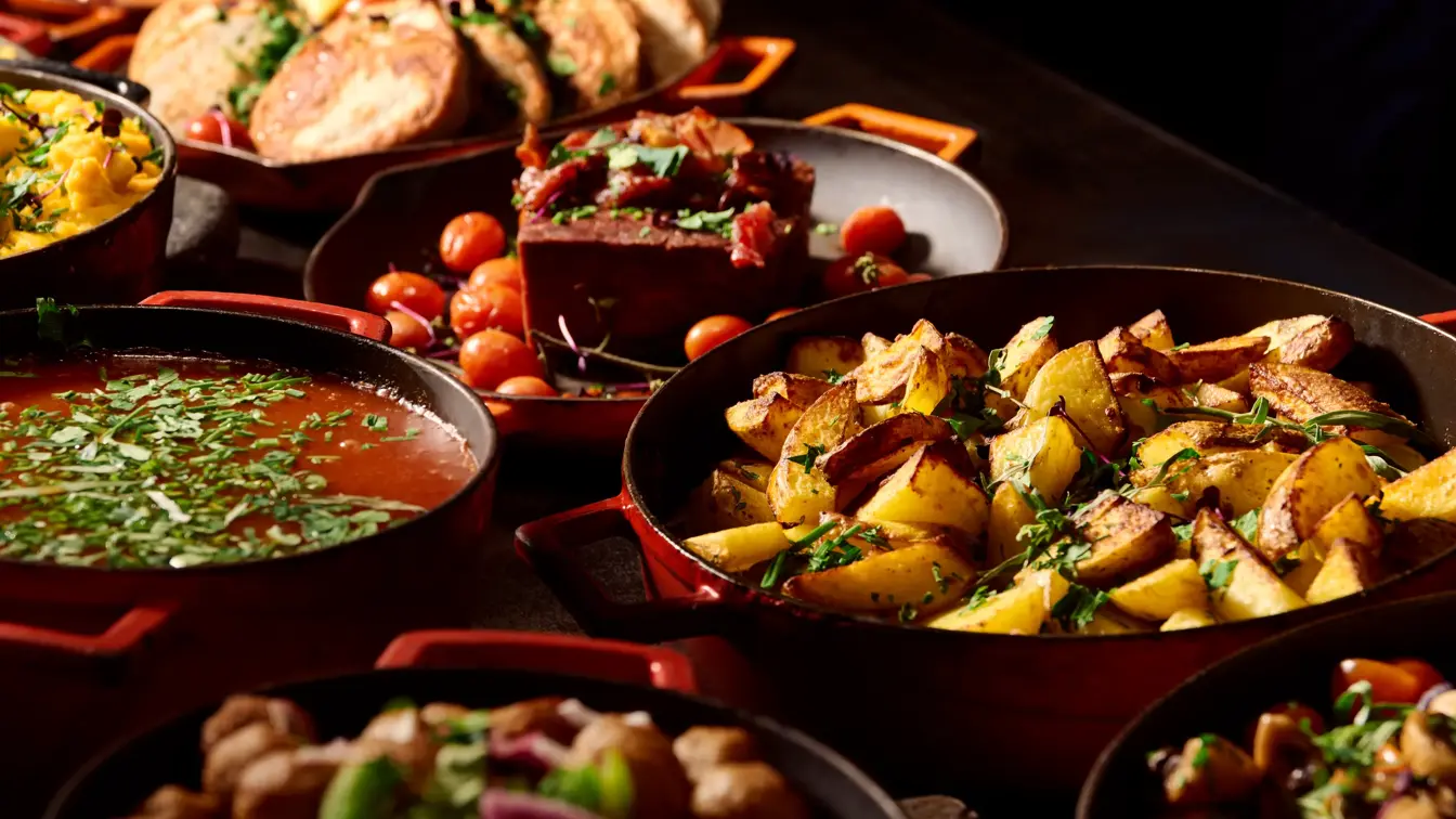 Nærbilde av en rekke runde, røde støpejernspanner fylt med ulike varme retter står på en frokostbuffet.