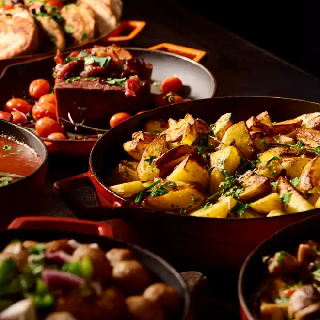 Nærbilde av en rekke runde, røde støpejernspanner fylt med ulike varme retter står på en frokostbuffet.