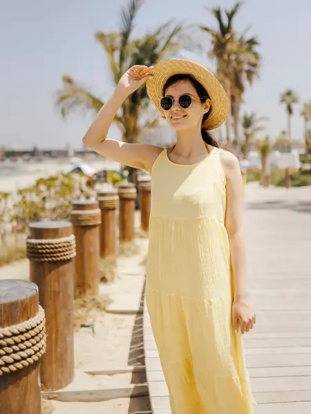 Dame med smørgul kjole, stråhatt og solbriller poserer på brygge ved strand 