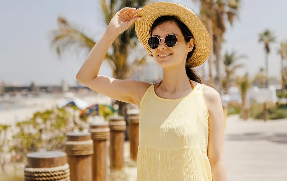 Dame med smørgul kjole, stråhatt og solbriller poserer på brygge ved strand 