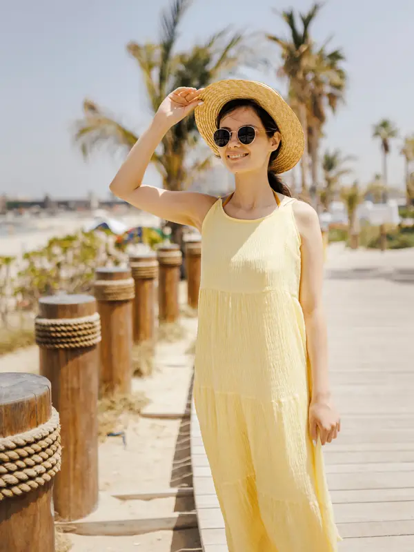 Dame med smørgul kjole, stråhatt og solbriller poserer på brygge ved strand 