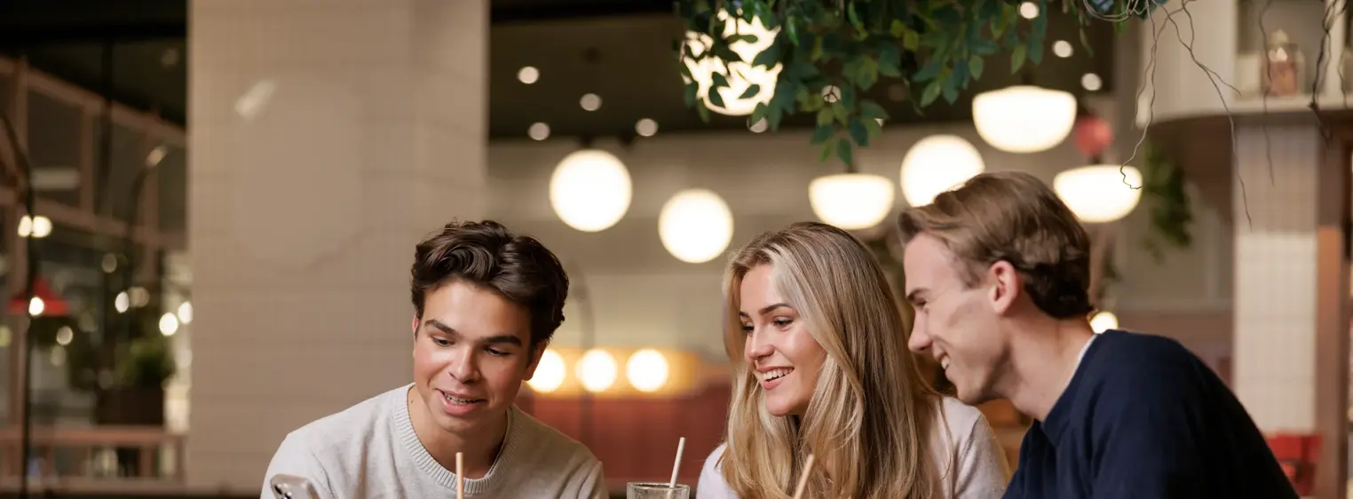 To gutter og en jente som sitter ved et bord i en kafé og ser på mobilen den ene gutten holder i hånden