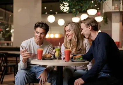 To gutter og en jente som sitter ved et bord i en kafé og ser på mobilen den ene gutten holder i hånden